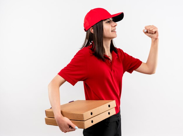 Młoda dziewczyna dostawy w czerwonym mundurze i czapce trzyma pudełka po pizzy zaciskając pięść, patrząc pewnie stojąc nad białą ścianą