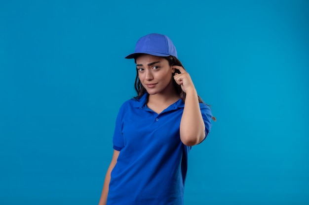 Młoda dziewczyna dostawy w niebieskim mundurze i czapce drapiąca głowę patrząc na kamery ze zdezorientowanym wyrazem twarzy mającej wątpliwości stojąc na niebieskim tle