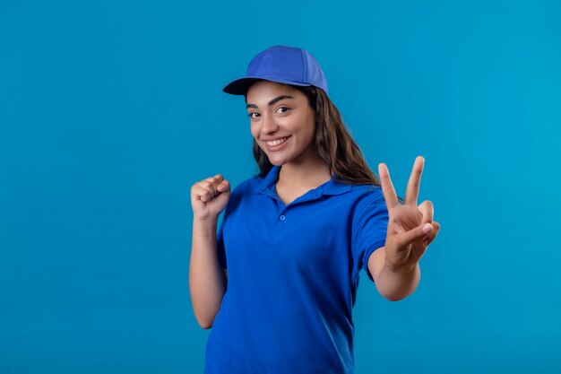 Młoda dziewczyna dostawy w niebieskim mundurze i czapce stojącej z zaciśniętą pięścią pokazującą znak zwycięstwa lub numer dwa uśmiechnięty wesoło, szczęśliwy i pozytywny stojąc na niebieskim tle