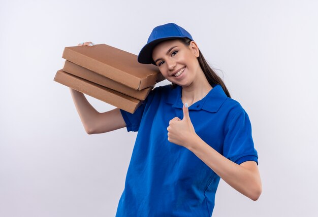 Młoda dziewczyna dostawy w niebieskim mundurze i czapce trzyma stos pudełek po pizzy patrząc na kamery uśmiechnięty radośnie pokazując kciuki do góry