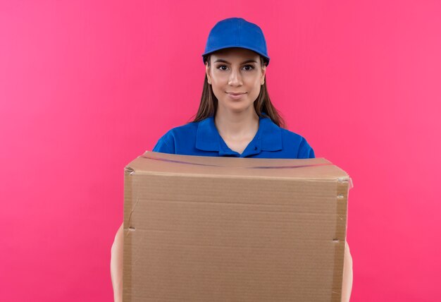 Młoda dziewczyna dostawy w niebieskim mundurze i czapce, trzymając duży pakiet pudełkowy patrząc na kamery z poważnym wyrazem pewności