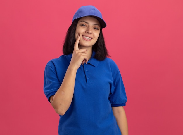 Młoda dziewczyna dostawy w niebieskim mundurze i czapce, uśmiechając się z radosną buźką z palcem na jej policzku stojąc na różowej ścianie