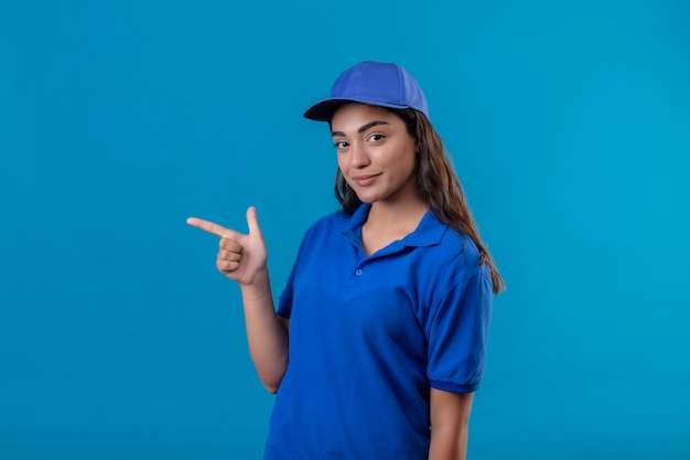 Młoda dziewczyna dostawy w niebieskim mundurze i czapce, wskazując palcem w bok, uśmiechnięta pewnie szczęśliwa i pozytywna pozycja na niebieskim tle