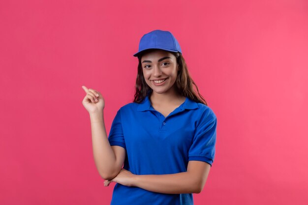 Młoda dziewczyna dostawy w niebieskim mundurze i czapce, wskazując palcem w bok, uśmiechnięta pewnie, szczęśliwa i pozytywna pozycja na różowym tle