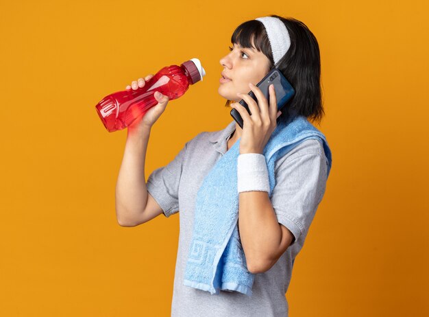Młoda dziewczyna fitness nosi opaskę z ręcznikiem na ramieniu, trzymając butelkę wody, wyglądając pewnie podczas rozmowy przez telefon komórkowy