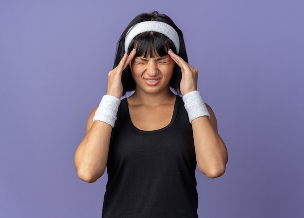 Bezpłatne zdjęcie młoda dziewczyna fitness nosząca opaskę, wyglądająca źle, dotykając głowy cierpiącej na ból głowy stojący na niebieskim tle