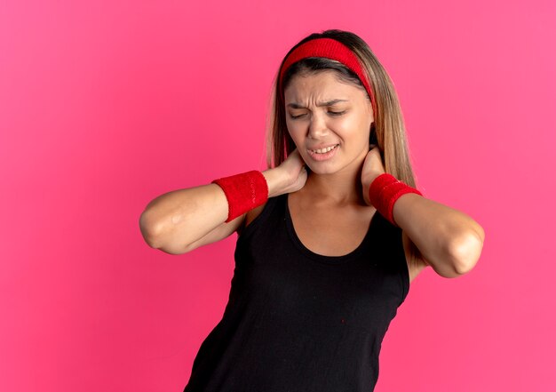 Młoda dziewczyna fitness w czarnej odzieży sportowej i czerwonej opasce dotyka jej szyi, patrząc na zły ból na różowo