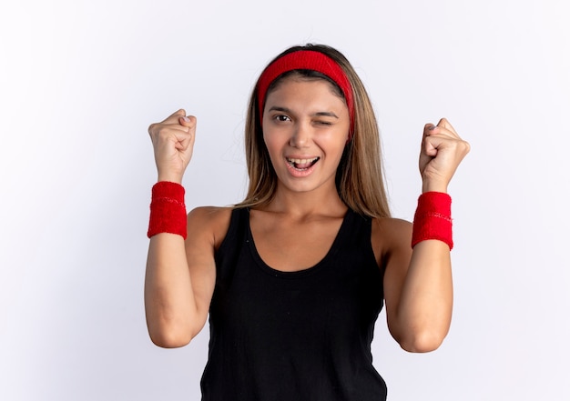 Młoda dziewczyna fitness w czarnej odzieży sportowej i czerwonej opasce lookign zaciskając pięści, mrugając i uśmiechając się stojąc nad białą ścianą
