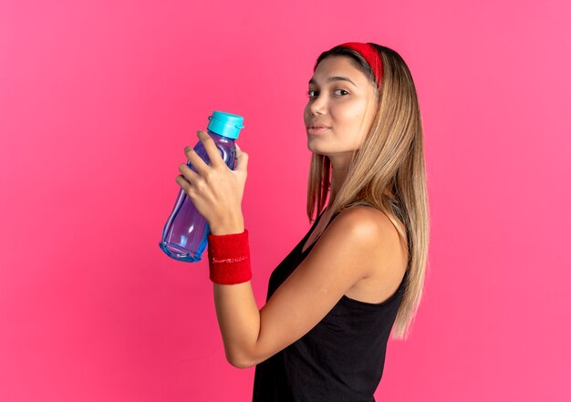 Młoda dziewczyna fitness w czarnej odzieży sportowej i czerwonej opasce trzyma butelkę wody, uśmiechając się pewnie na różowo
