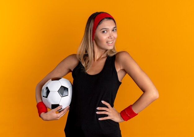 Młoda dziewczyna fitness w czarnej odzieży sportowej i czerwonej opasce trzyma piłkę nożną patrząc pewnie z uśmiechem stojąc na pomarańczowej ścianie