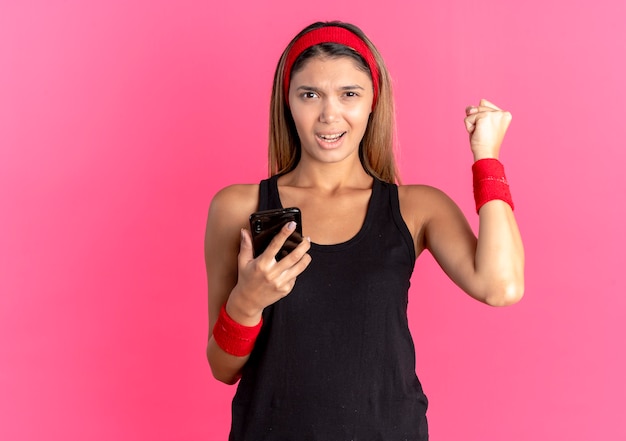 Młoda dziewczyna fitness w czarnej odzieży sportowej i czerwonej opasce trzyma smartfon zaciskając pięść szczęśliwy i podekscytowany stojąc nad różową ścianą