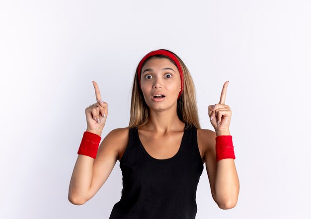 Bezpłatne zdjęcie młoda dziewczyna fitness w czarnej odzieży sportowej i czerwonej opasce wygląda zaskoczony, wskazując palcami wskazującymi, stojąc na białej ścianie
