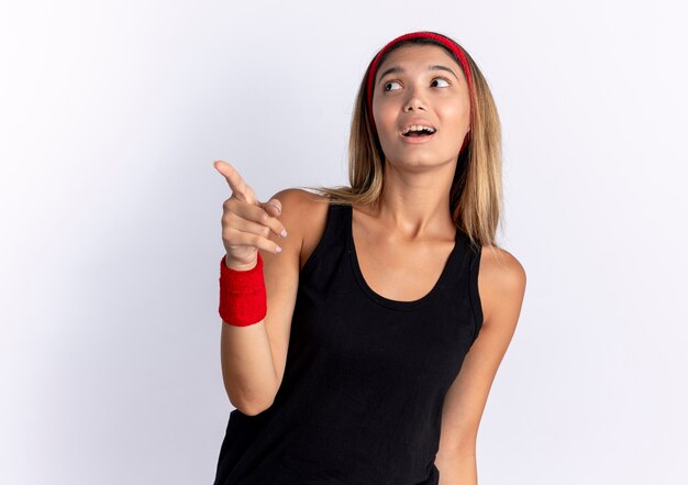 Młoda dziewczyna fitness w czarnej odzieży sportowej i czerwonej opasce wyglądającej na zdezorientowanego wskazującego palcem na coś przestraszonego stojącego na białej ścianie