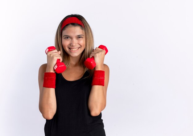 Młoda dziewczyna fitness w czarnej odzieży sportowej i czerwonej opasce z hantlami, uśmiechając się wesoło stojąc na białej ścianie