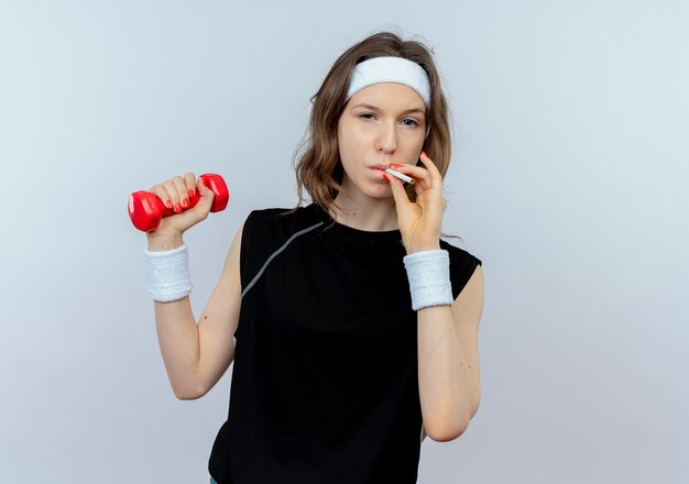 Młoda dziewczyna fitness w czarnej odzieży sportowej z pałąkiem na głowę, ćwicząc z hantlami i paleniem papierosów koncepcja złego nawyku, stojąc na białej ścianie