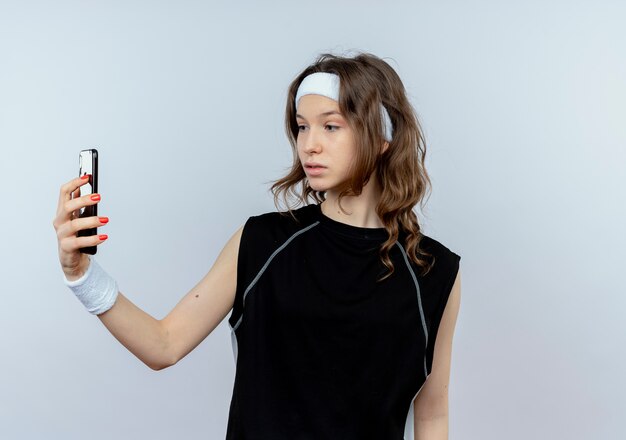 Młoda dziewczyna fitness w czarnej odzieży sportowej z pałąkiem na głowę patrząc na ekran smartfona z sceptycznym wyrazem twarzy stojącej na białej ścianie