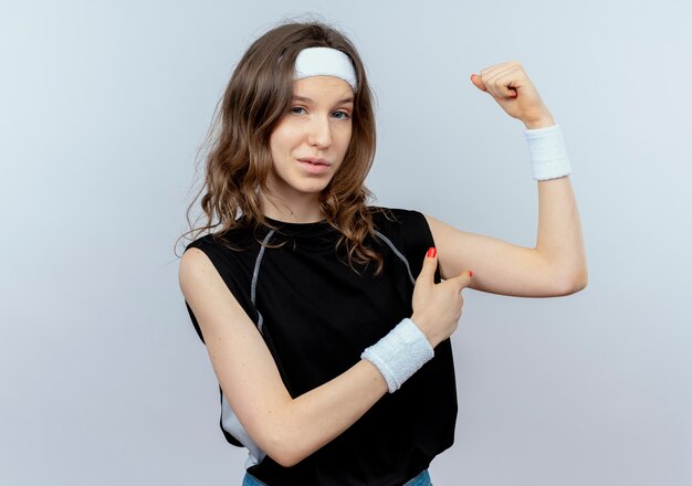 Młoda dziewczyna fitness w czarnej odzieży sportowej z pałąkiem na głowę podnoszącą rękę pokazującą biceps lookign pewnie stojąc na białej ścianie