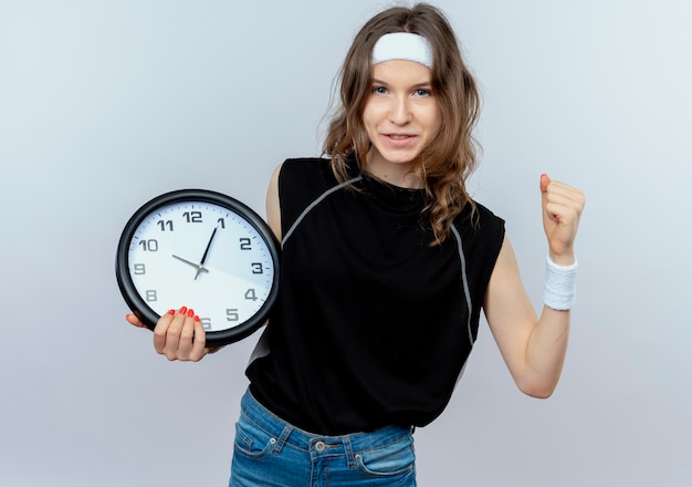 Młoda dziewczyna fitness w czarnej odzieży sportowej z pałąkiem na głowę trzyma zegar ścienny uśmiechnięta pewnie, zaciskając pięść stojącą nad białą ścianą