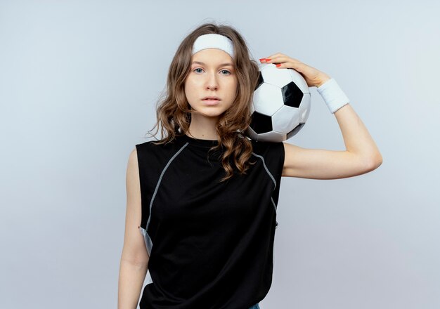 Młoda dziewczyna fitness w czarnej odzieży sportowej z pałąkiem na głowę trzymając piłkę nożną z poważną twarzą stojącą na białej ścianie