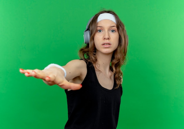 Bezpłatne zdjęcie młoda dziewczyna fitness w czarnej odzieży sportowej z pałąkiem na głowę ze słuchawkami uspokajający gest ręką stojącą na zielonej ścianie