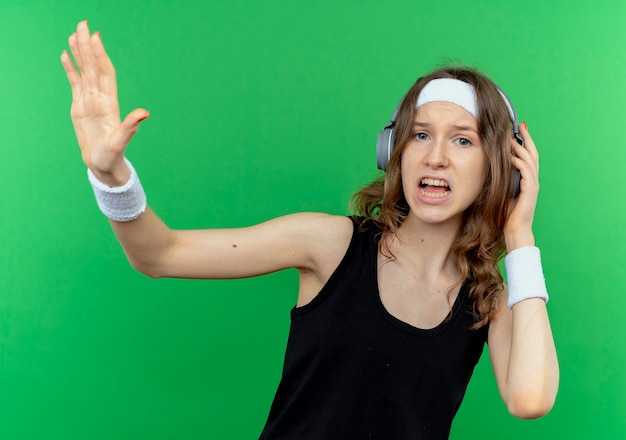 Młoda dziewczyna fitness w czarnej odzieży sportowej z pałąkiem na głowę ze słuchawkami wyglądająca na zdezorientowaną i niezadowoloną, robiąc znak stopu ręką stojącą nad zieloną ścianą