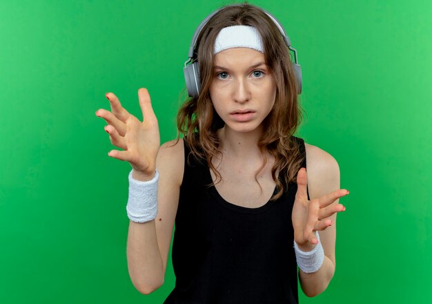 Młoda dziewczyna fitness w czarnej odzieży sportowej z pałąkiem na głowę ze słuchawkami z poważną twarzą gestykuluje rękami stojącymi nad zieloną ścianą