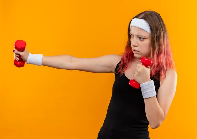 Młoda dziewczyna fitness w odzieży sportowej z hantlami robi ćwiczenia patrząc pewnie stojąc na pomarańczowej ścianie