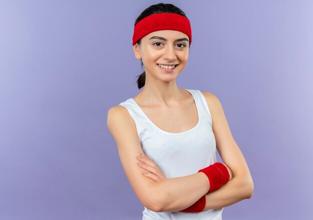 Młoda dziewczyna fitness w odzieży sportowej z pałąkiem na głowę z pewnym uśmiechem stojącym nad fioletową ścianą