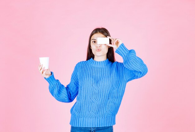młoda dziewczyna model trzyma kartę i plastikowy kubek na różowej ścianie.