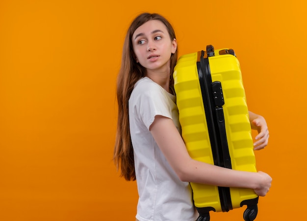 Młoda dziewczyna podróżnika, trzymając walizkę, patrząc na lewą stronę na odizolowanej pomarańczowej ścianie z miejsca na kopię