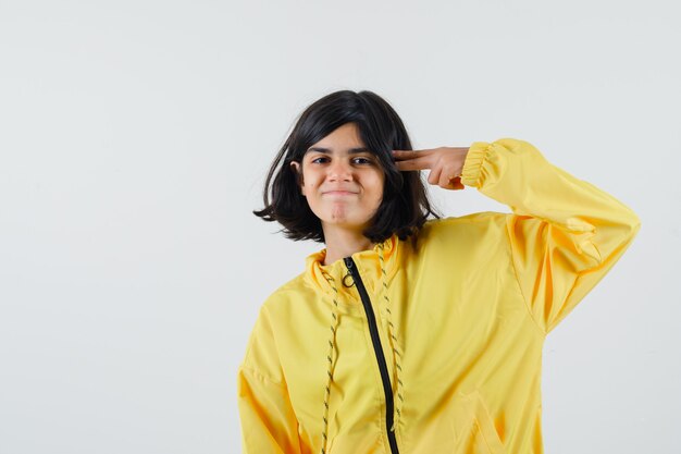 Młoda dziewczyna pokazuje gest pistoletu w pobliżu głowy w żółtej kurtce bombowej i wygląda na szczęśliwego