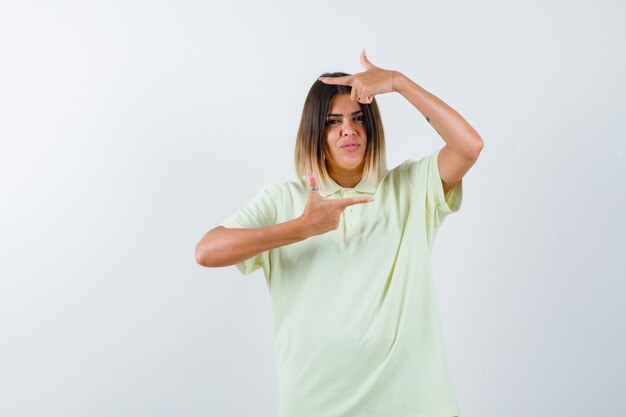 Młoda dziewczyna pokazuje gest ramy w koszulce i wygląda poważnie. przedni widok.