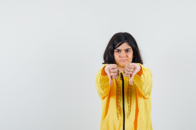 Młoda dziewczyna pokazuje kciuki w dół obiema rękami w żółtej bomberce i wygląda na niezadowoloną.
