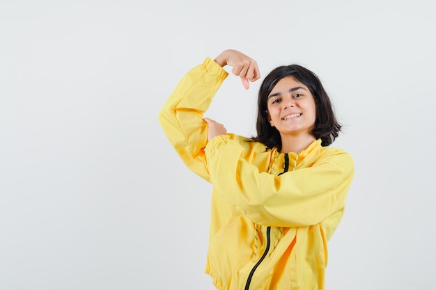 Młoda dziewczyna pokazuje mięśnie w żółtej bomberce i wygląda szczęśliwie.
