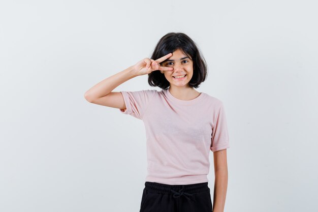 Młoda dziewczyna pokazuje znak v na oku w różowej koszulce i czarnych spodniach i wygląda uroczo