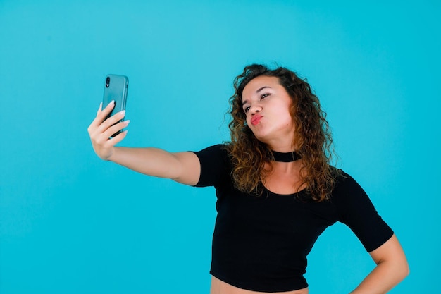 Młoda dziewczyna robi selfie telefonem, pokazując mimikę pocałunku na niebieskim tle