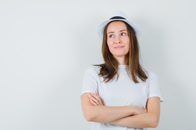 Młoda dziewczyna stoi ze skrzyżowanymi rękami w białym kapeluszu t-shirt i marzycielski wygląd