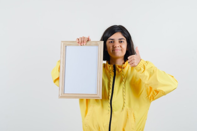 Młoda dziewczyna trzyma białą tablicę i pokazuje kciuk w żółtej kurtce bombowej i wygląda na szczęśliwego