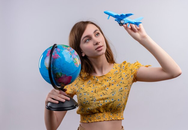 Młoda dziewczyna trzyma kulę ziemską i model samolotu i patrząc na model samolotu na odizolowanej białej ścianie