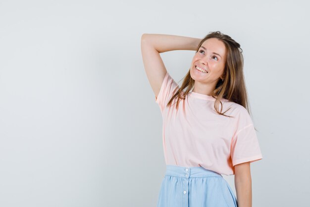 Młoda dziewczyna trzyma podniesioną rękę na głowie w t-shirt, spódnicy i patrząc z nadzieją, widok z przodu.