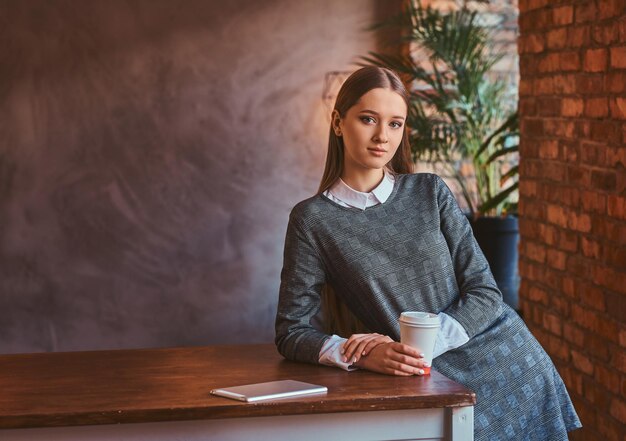 Młoda dziewczyna ubrana w elegancką szarą sukienkę trzyma filiżankę kawy na wynos, patrząc w kamerę, opierając się na stole w pokoju z wnętrzem na poddaszu.