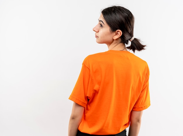 Młoda dziewczyna ubrana w pomarańczowy t-shirt mimo jej pleców patrząc na bok na białej ścianie