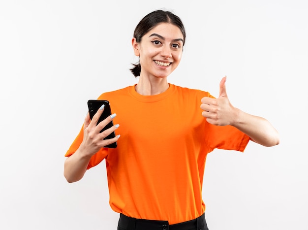 Młoda dziewczyna ubrana w pomarańczowy t-shirt trzymając smartfon pokazując kciuki do góry uśmiechnięty wesoło stojąc na białej ścianie