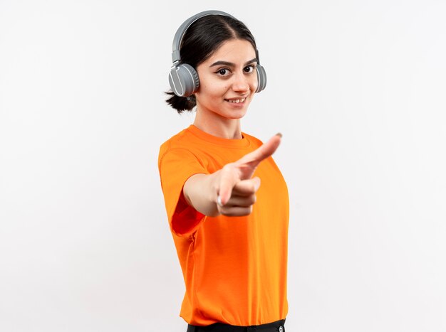 Młoda dziewczyna ubrana w pomarańczowy t-shirt ze słuchawkami szczęśliwa i pozytywna, ciesząc się swoją ulubioną muzyką, uśmiechając się, wskazując palcem wskazującym stojącym na białej ścianie