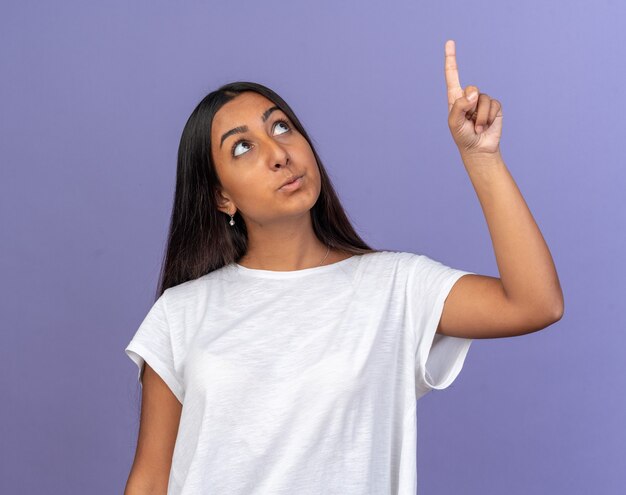 Młoda dziewczyna w białej koszulce patrząca w górę zaintrygowana, wskazując palcem wskazującym na coś stojącego nad niebieskim