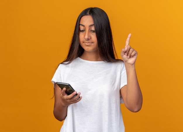 Młoda dziewczyna w białej koszulce trzymając smartfon patrząc na to z uśmiechem na inteligentnej twarzy pokazując palec wskazujący mający nowy pomysł stojący na pomarańczowym tle