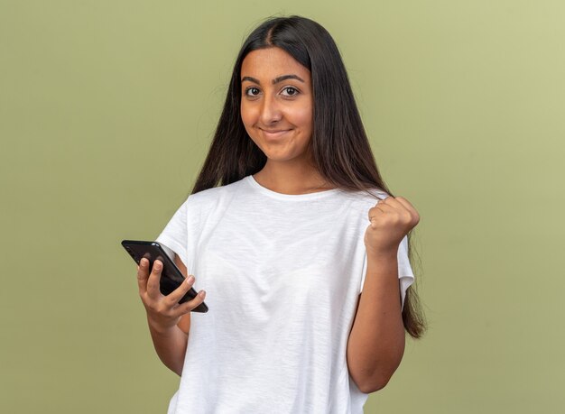 Młoda dziewczyna w białej koszulce trzymająca smartfona patrząca na kamerę szczęśliwa i podekscytowana zaciskająca pięść