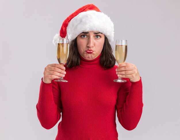 Młoda dziewczyna w czerwonym swetrze i czapce mikołaja trzymająca dwie szklanki szampana patrząc na kamerę zdezorientowana i niezadowolona stojąc na białym tle