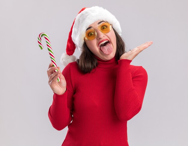 Młoda dziewczyna w czerwonym swetrze i santa hat w okularach trzymając kandyzowaną laskę szczęśliwa i wesoła wystający język stojący na białym tle