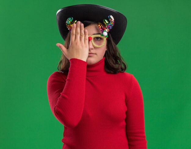 Młoda dziewczyna w czerwonym swetrze na sobie śmieszne okulary i czarny kapelusz zakrywający jedno oko ręką